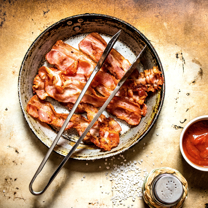 A pan with four slices of grass fed bacon and a pair of tongs.