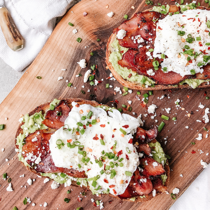 Two slices of avocado toast topped with bacon and a poached egg, dressed with green onion and pepper flakes on a wooden cutting board.