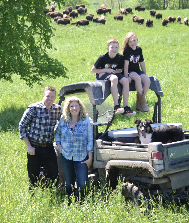 Two farmers leaning against a truck. Their two children sit on top of it.