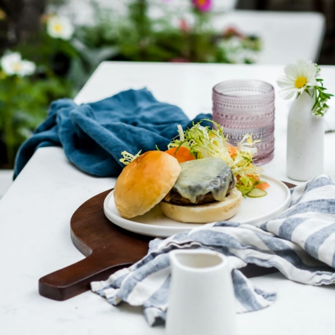 A grass fed burger on a bun with salad on the side. The table setting is a very white patio table dressed with cloth napkins and a daisy in a bottle.