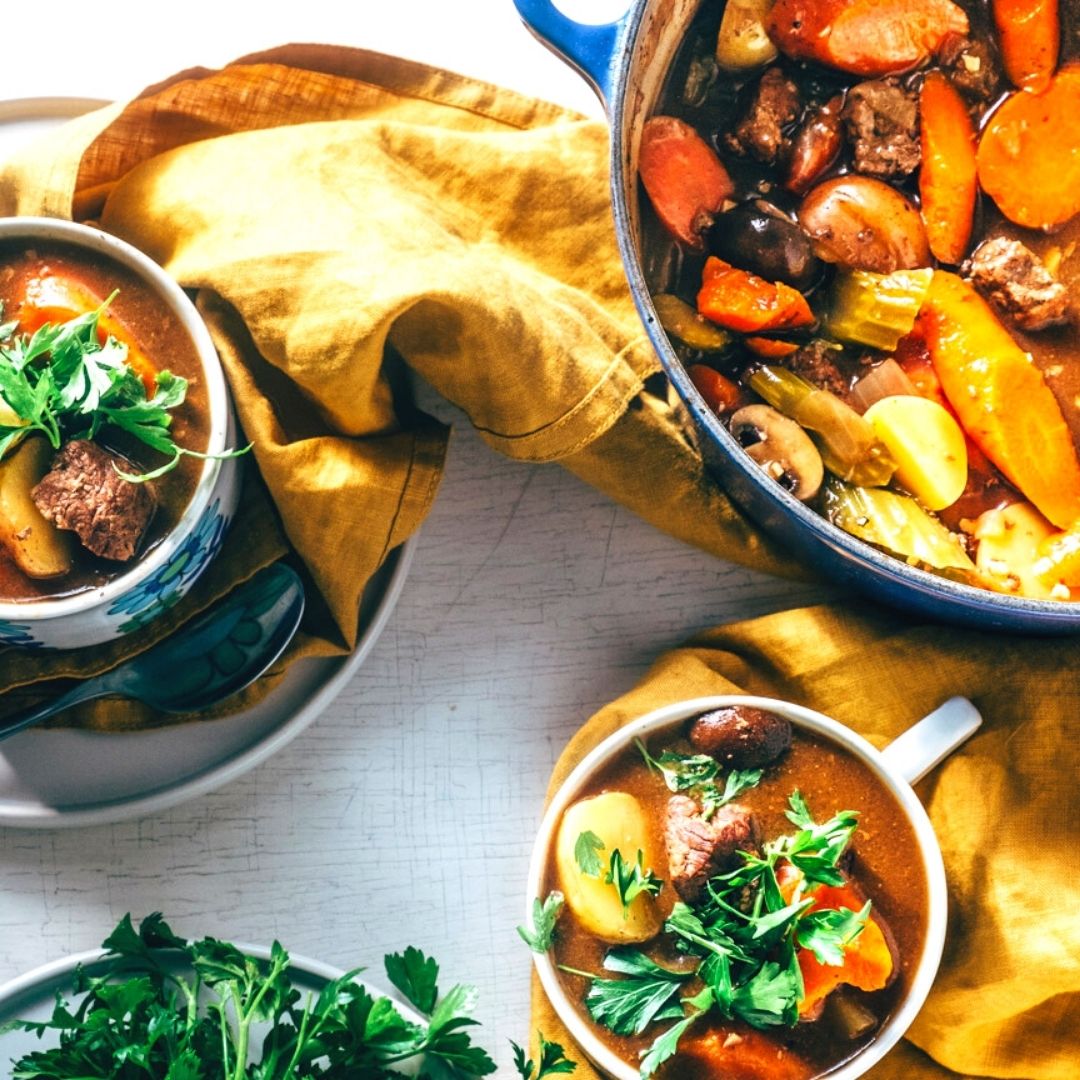 A big pot of beef stew with carrots and celery. Two cups filled with stew, topped with parsley.
