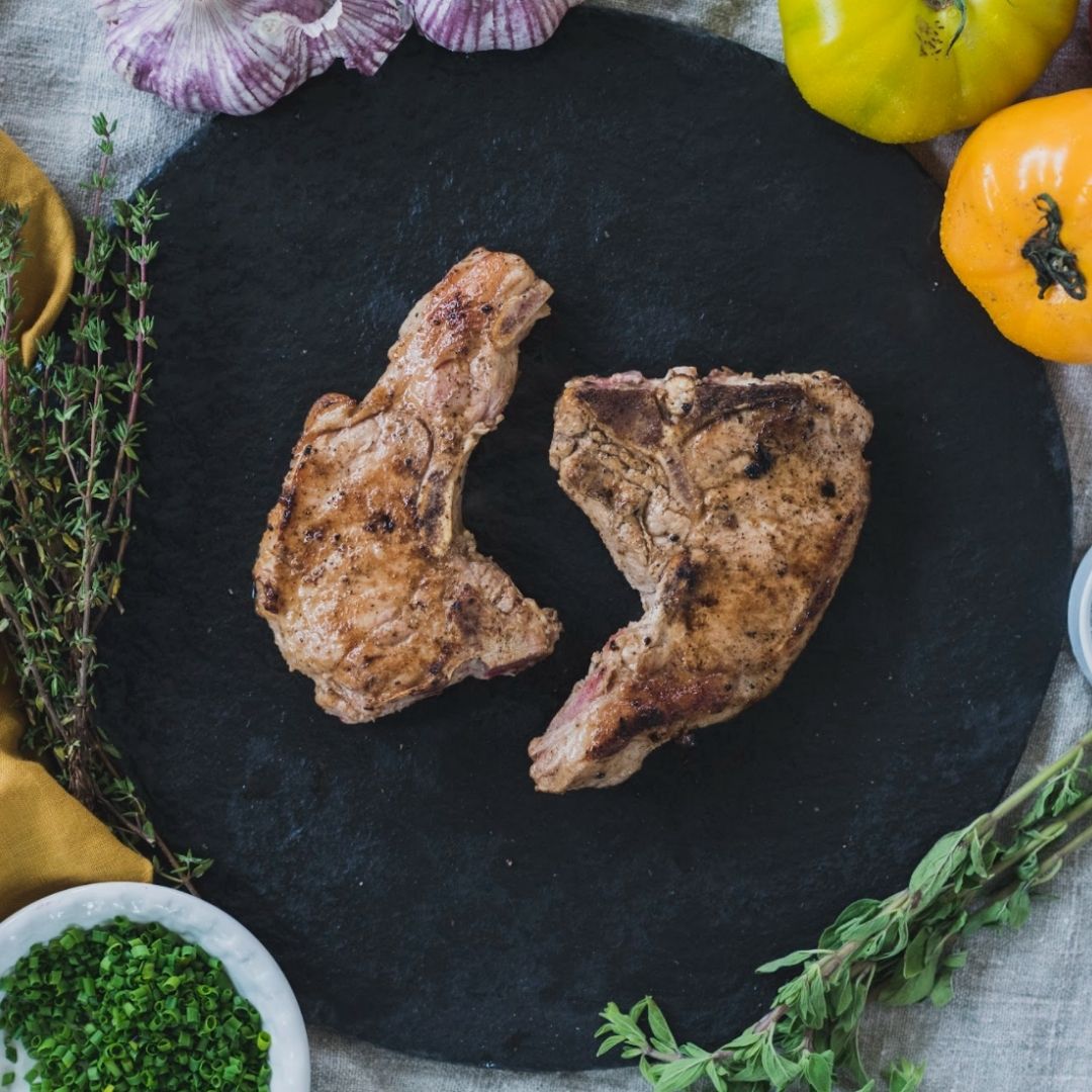 Two pasture raised pork chops in the centre of a black plate, surrounded by greens and vegetables.