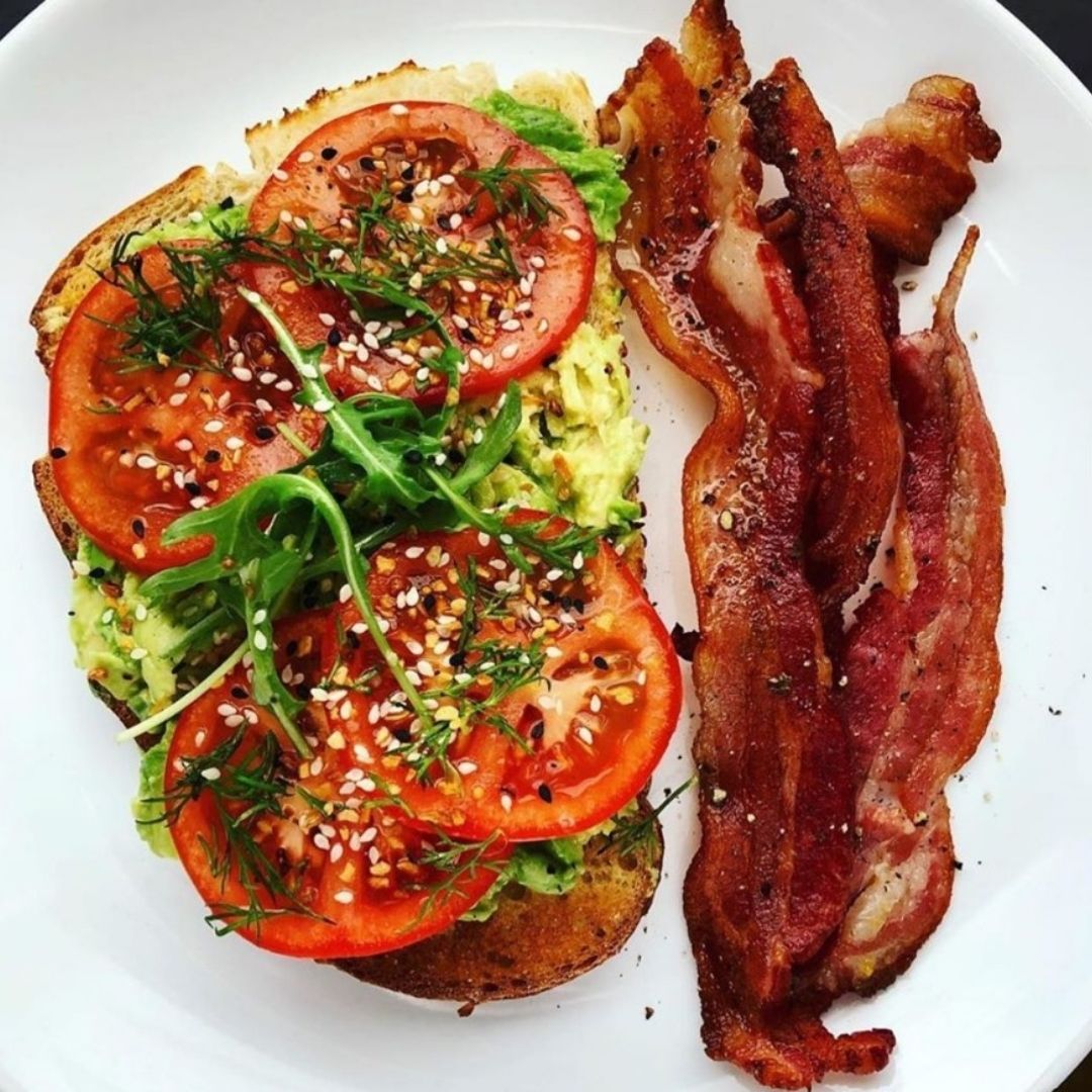 Avocado toast topped with tomatoes and arugula, with a side of pasture raised bacon.