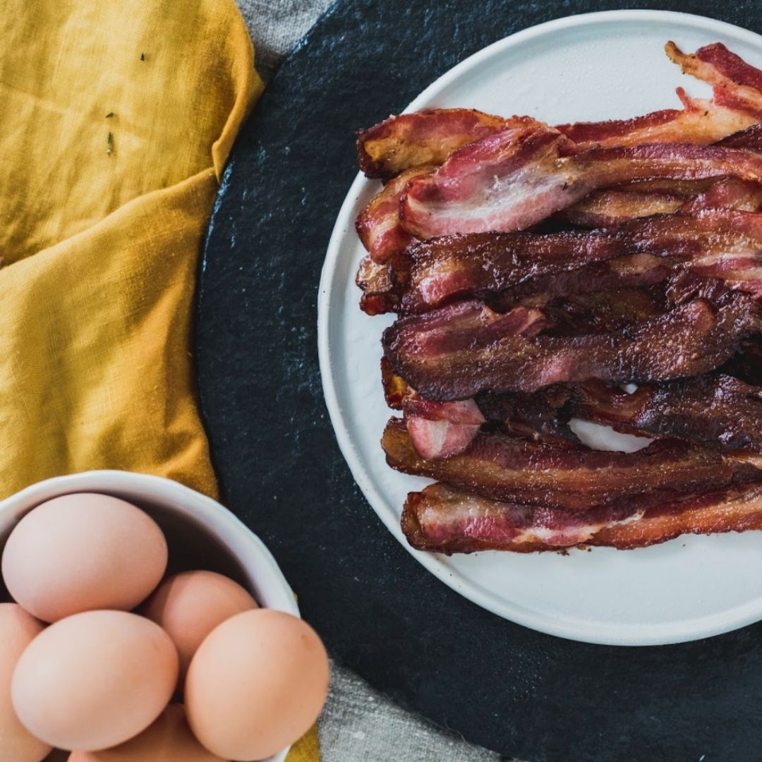About ten strips of bacon on a white plate next to a bowl of brown eggs and a yellow cloth napkin.