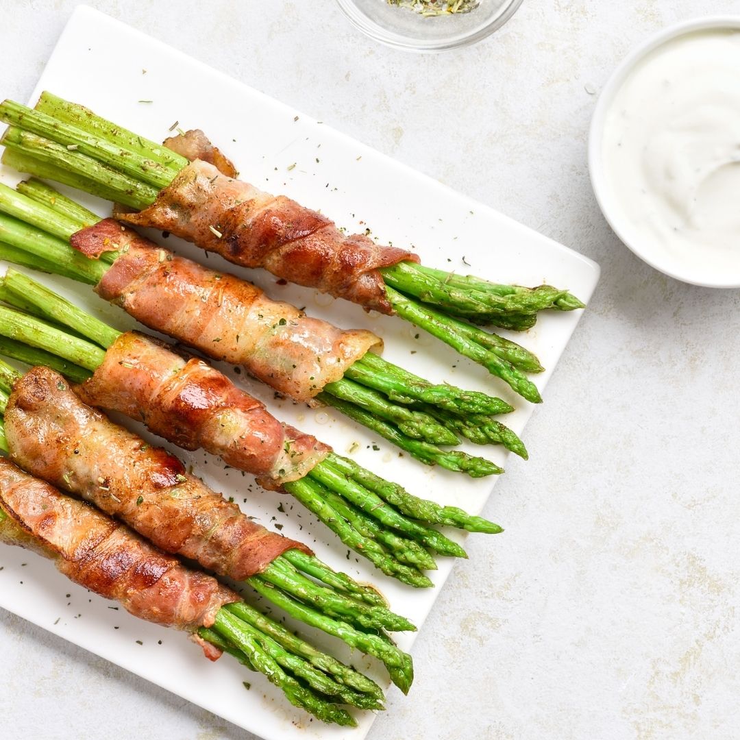 Four bunches of bacon wrapped asparagus on a square white plate. A small cup of white dipping sauce can be seen next to the plate.