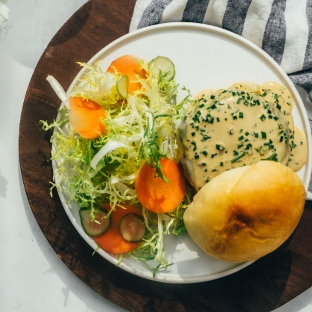 A top-down view of a white plate with a cheesy hamburger, the top bun off to the side, and a bright, green salad next to it.