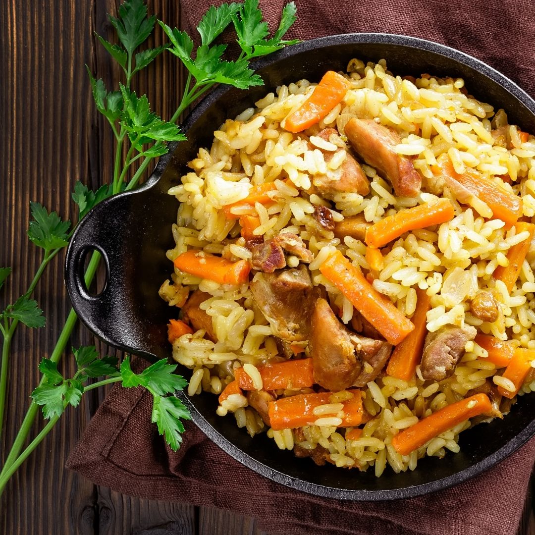 A black bowl filled with brown rice, shredded chicken and sliced carrots. It is on a wooden board next to a sprig of parsley.