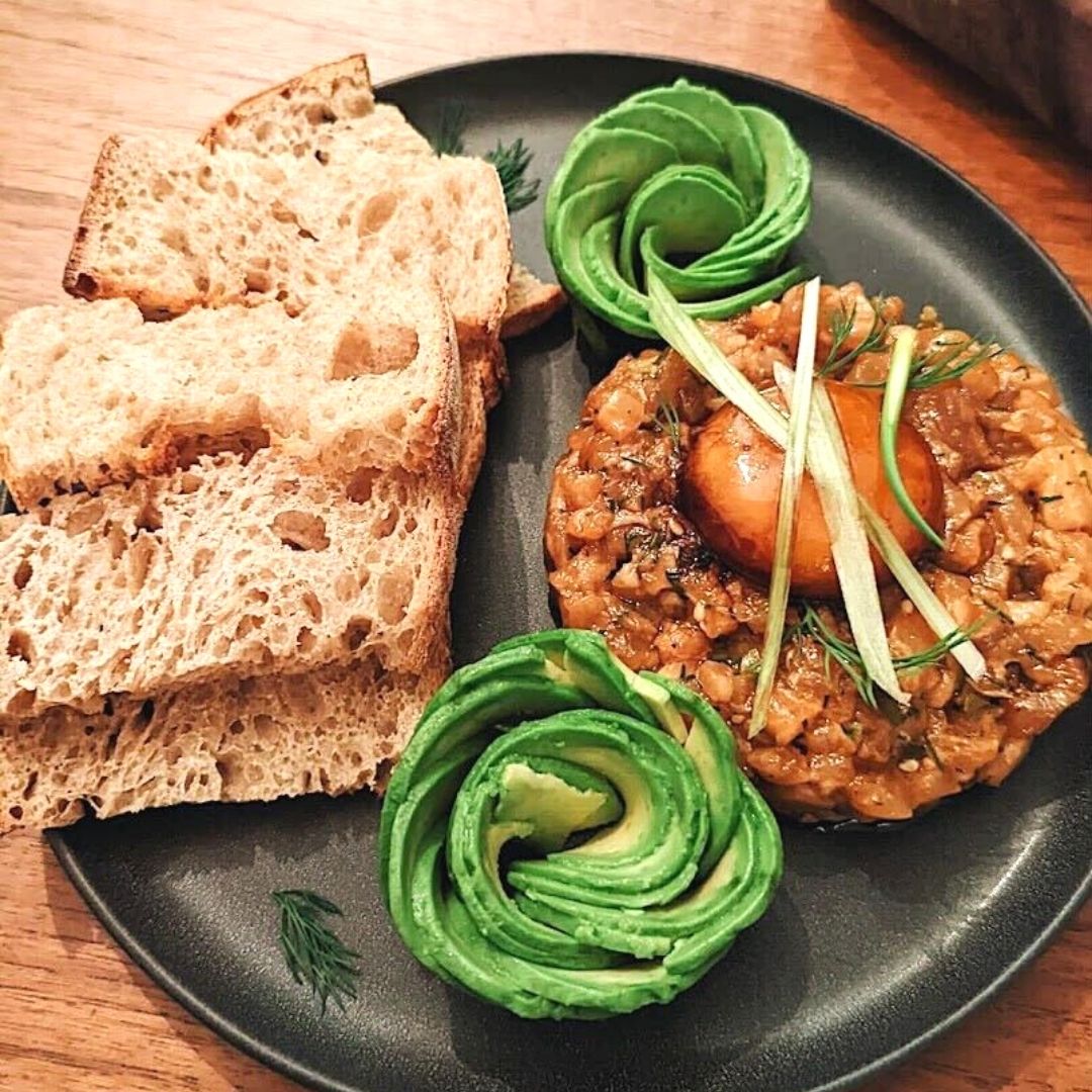 Trout tartare topped with egg yolk plated next to thin slices of bread and avocado slices arranged in two elegant swirls.
