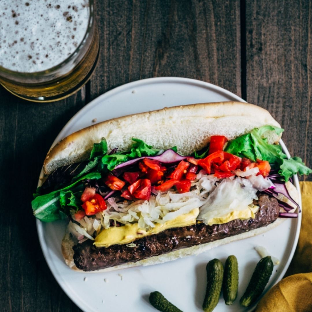 Pasture raised sausage on a bun with lettuce, tomato, onions and cheese. There are pickles on the plate with the bun, and a beer next to the plate.