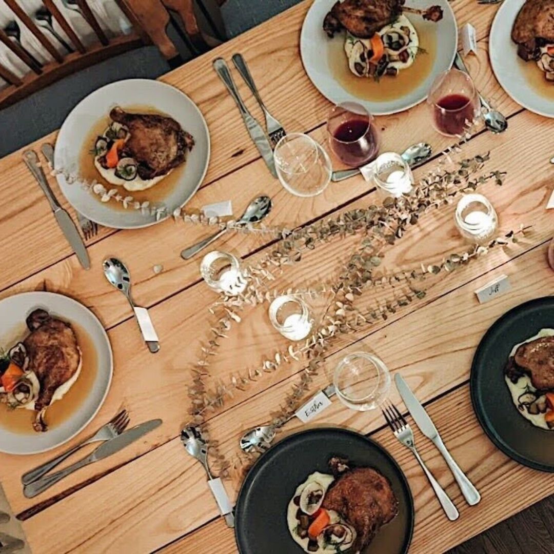 An overhead view of a wooden table set for six. There are candles, wine glasses, and the plates are filled with sophisticated poultry.