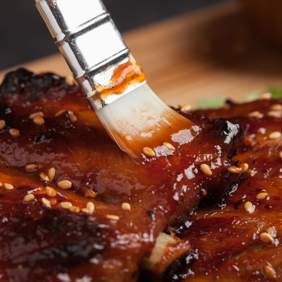 A white brush, coating a rack of ribs in barbecue sauce.