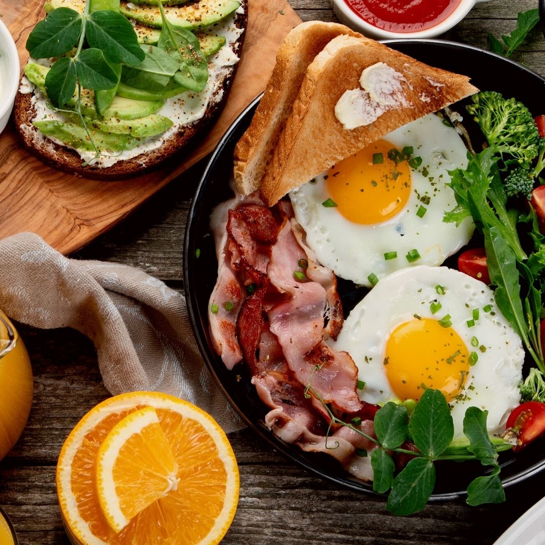 An overhead view of bacon, eggs, toast and veggies served in a black dish. The wooden table also has orange slices and a cutting board topped with avocado toast.