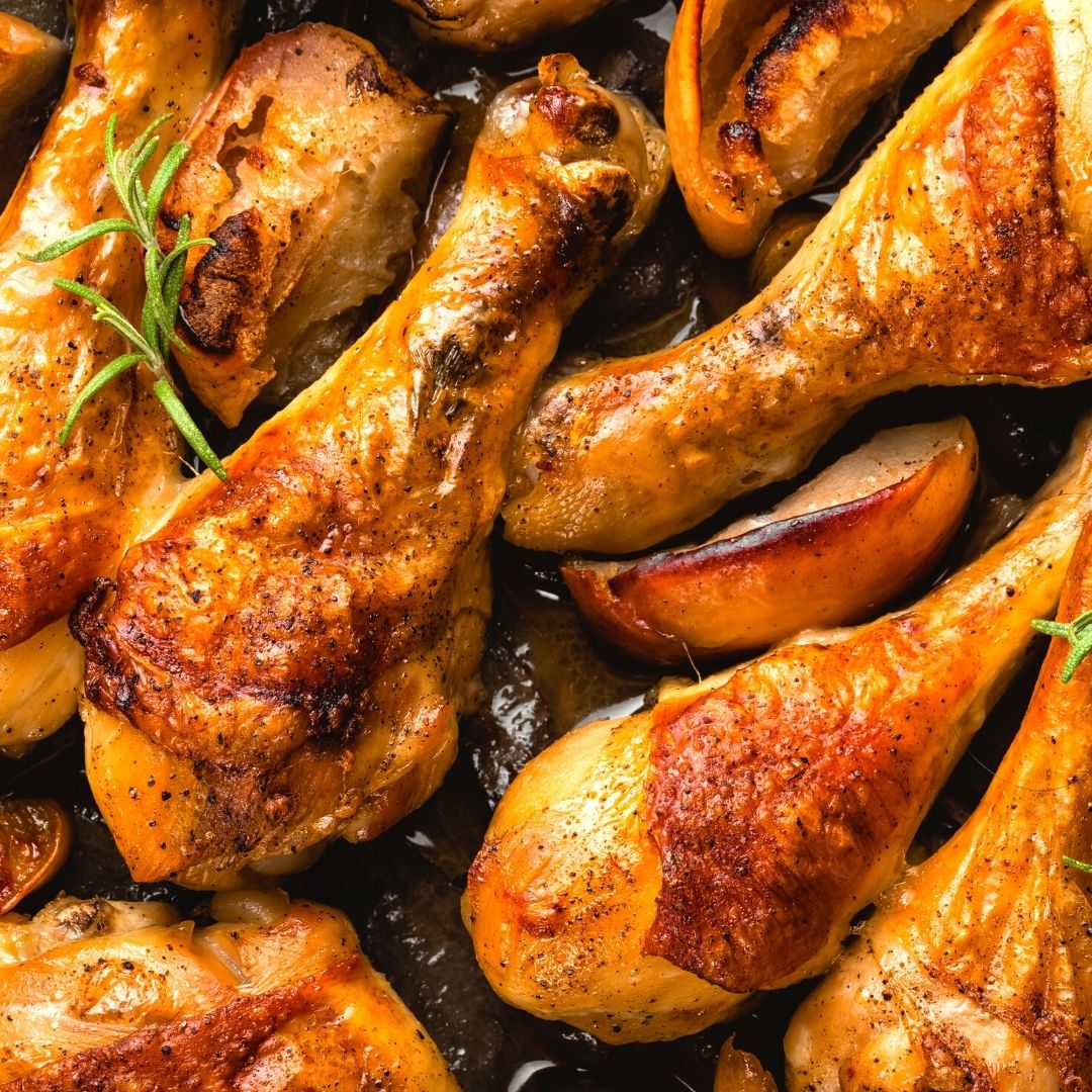 A pan of warm chicken drumsticks with crispy skin. A sprig of rosemary and some chopped potatoes are also in the pan.