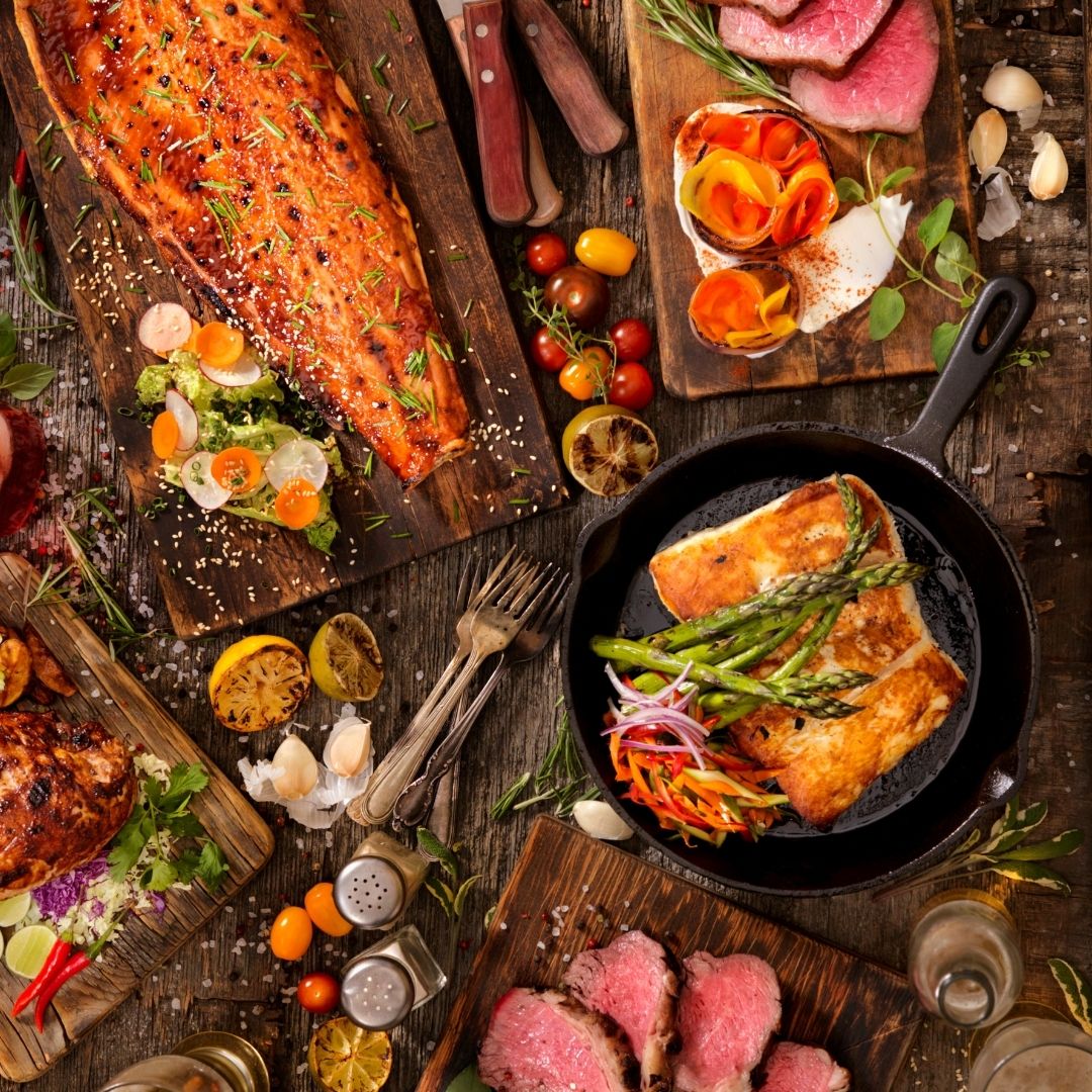 Overhead image of various fish and meat on a wooden table, on top of cutting boards, in cast iron pans, and prepared to be eaten. 