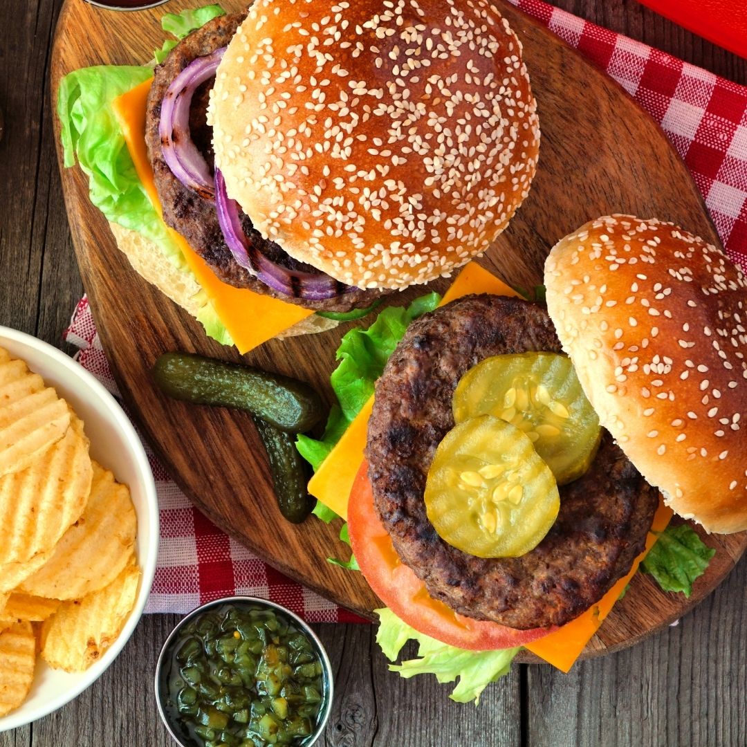 A cutting board with two loaded burgers on top. One bun is open, showcasing the pickles, cheese, lettuce and tomato inside. There is a small dish of relish to the side and a bowl with ridged potato chips.