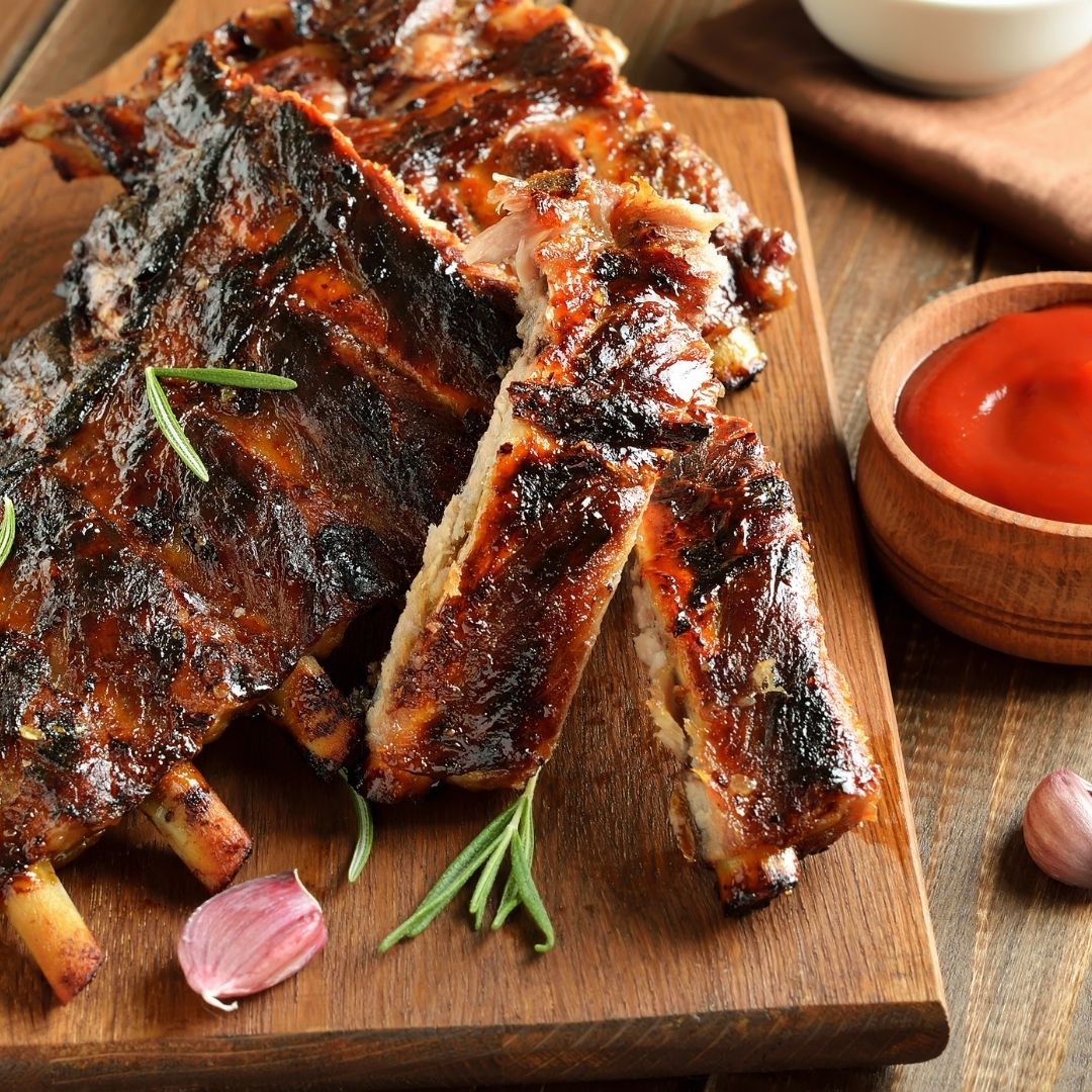 A succulent rack of pasture-raised ribs on a wooden cutting board. There is a small bowl of BBQ sauce next to the ribs. 