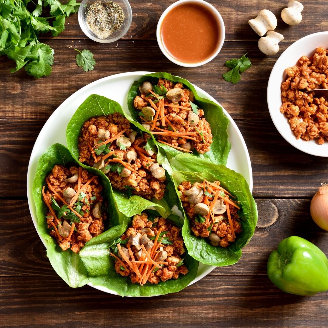 Five lettuce wraps filled with meat, herbs and vegetables on a white plate. The plate is on a wooden table, surrounded by vegetables, herbs, and other ingredients.