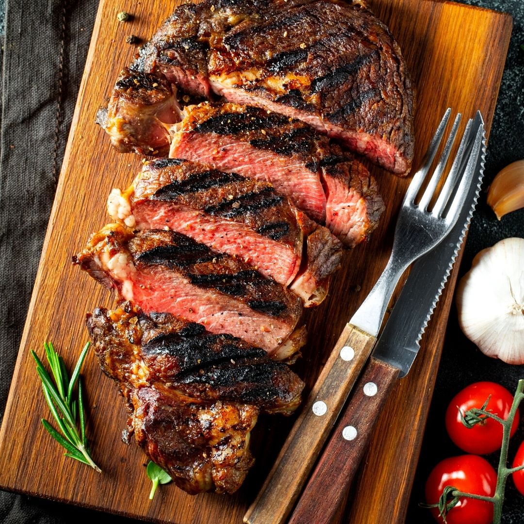 A medium-rare steak sliced on a wooden cutting board. There are tomatoes, garlic, herbs and a fork and knife next to the steak.