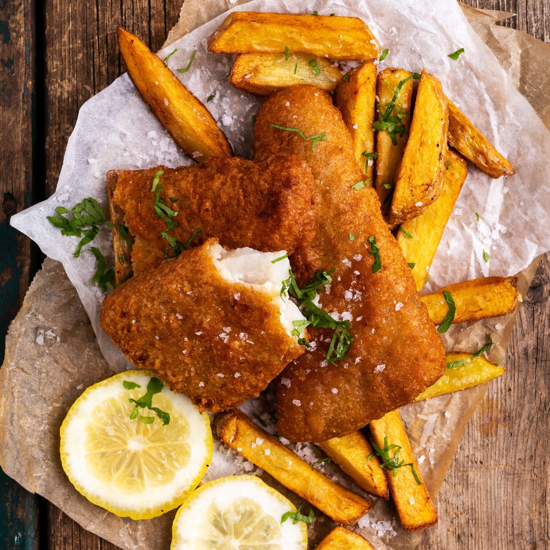 Three pieces of beer-battered fish fillet on a wooden table. They sit on top of crispy french fries and two slices of lemon.