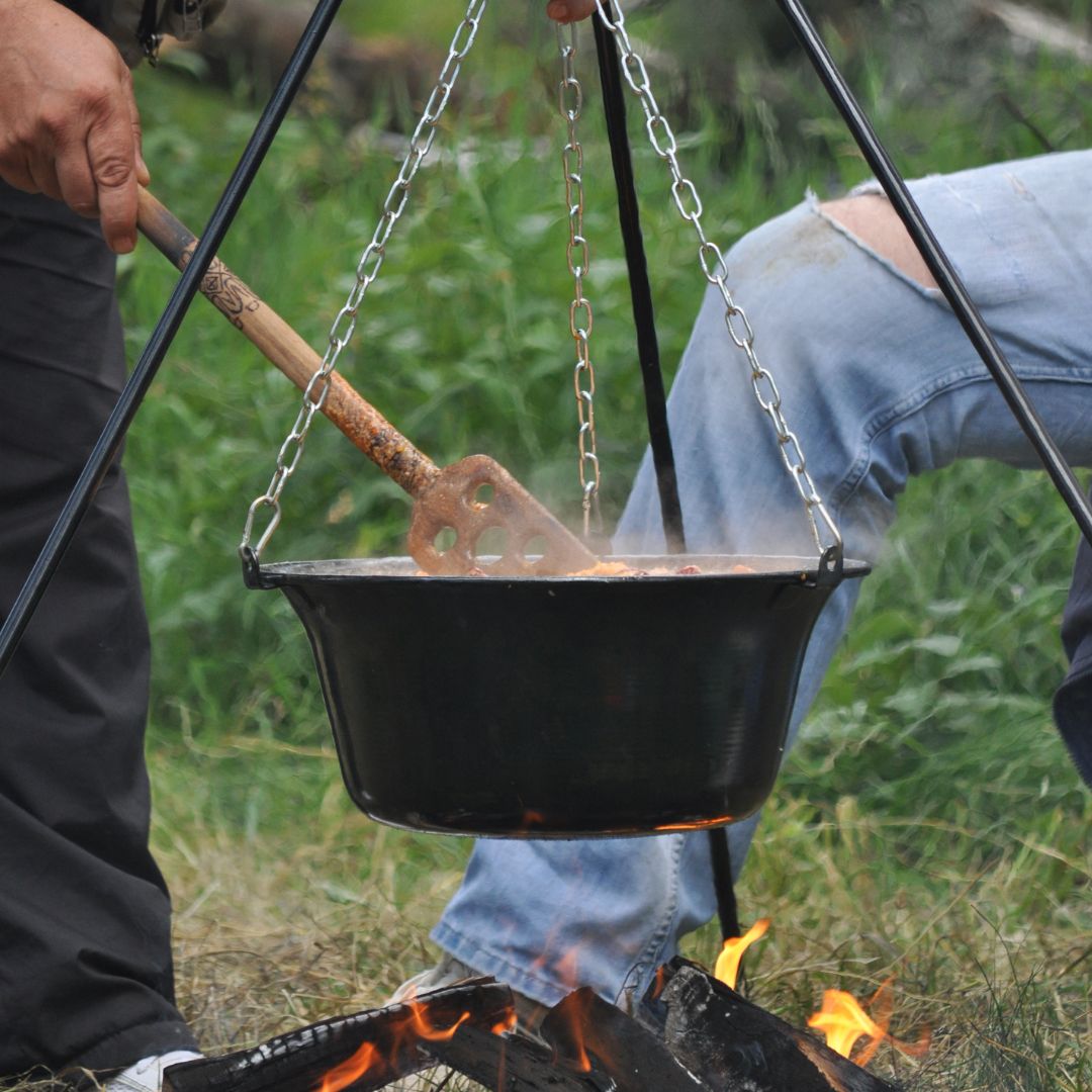 Two people sitting next to a campfire. A pot of food sitting over the fire, and one person is stirring the food.