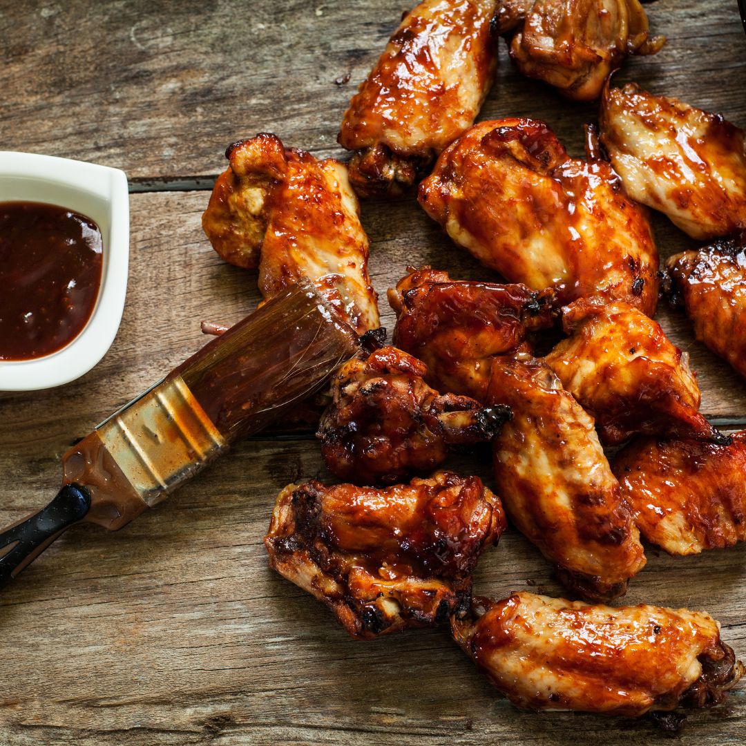 A pound of chicken wings spread on a wooden table. A BBQ sauce brush sits on top of the wings, and a cup of BBQ sauce is next to the wings.
