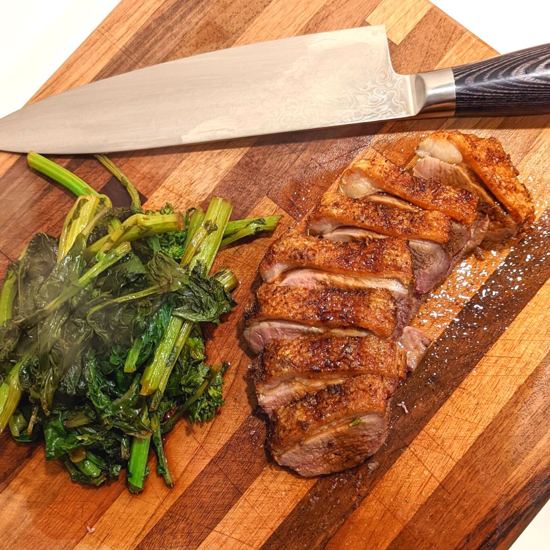 A cut of meat sliced thinly on a wooden cutting board, next to a pile of greens. A carving knife sits above them on the cutting board.