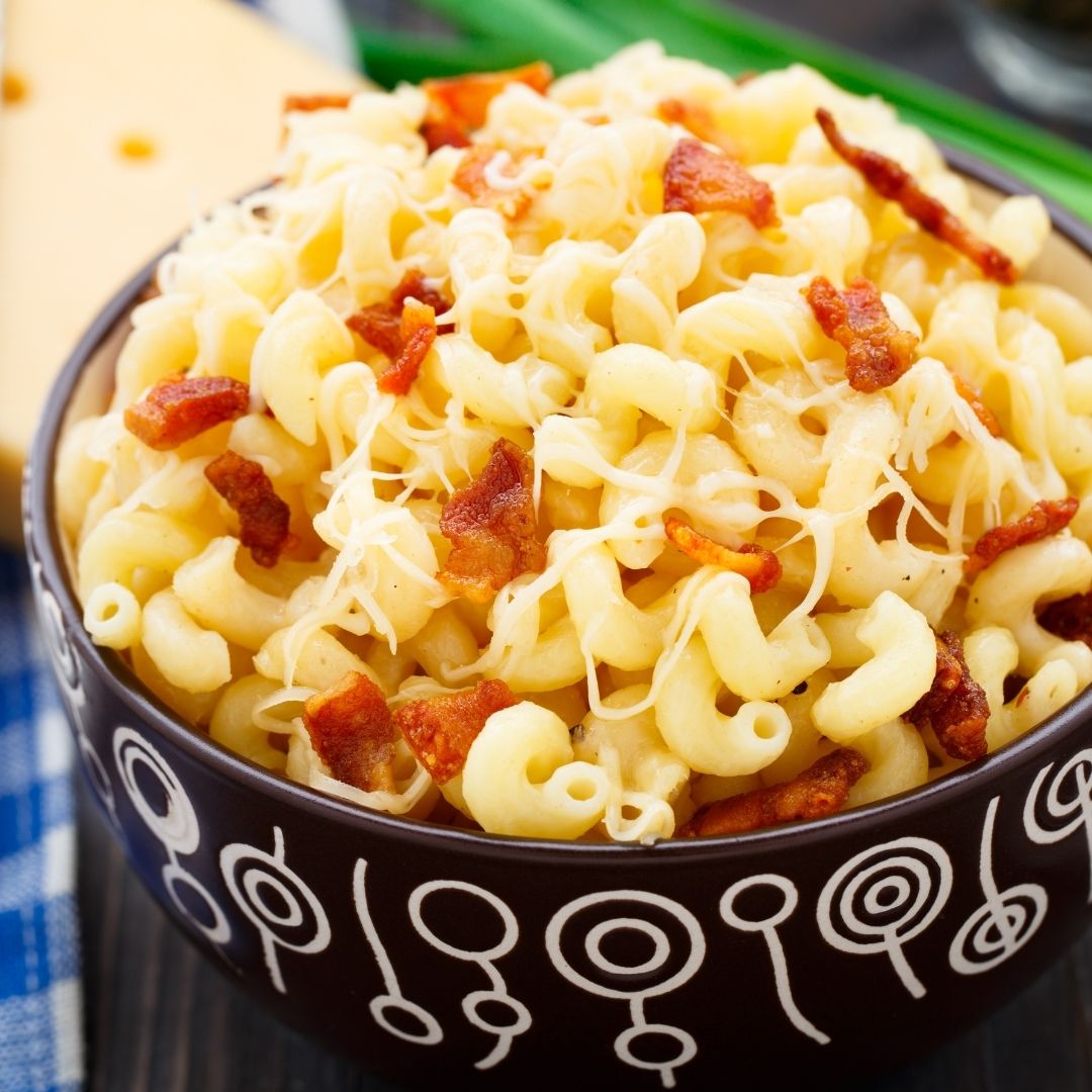A black bowl filled with macaroni and cheese with bacon. It sits on a blue checkered napkin with a block of cheese in the background.