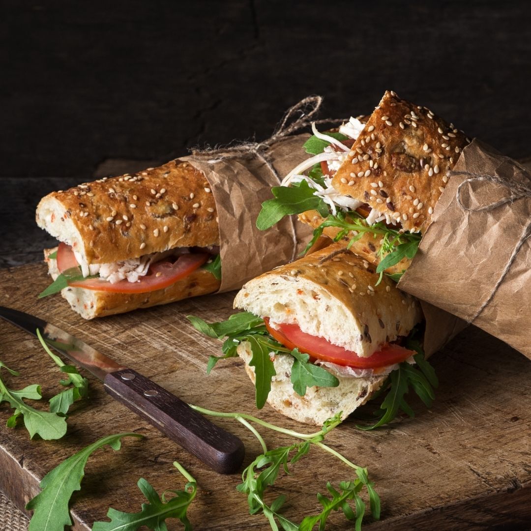 Three long sesame seed sandwiches topped with arugula, turkey and tomatoes next to a knife on a cutting board.