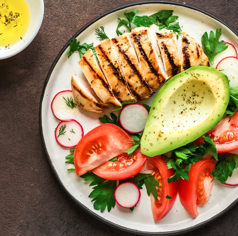An overhead view of a white plate topped with tomato, avocado, and grilled chicken breast.